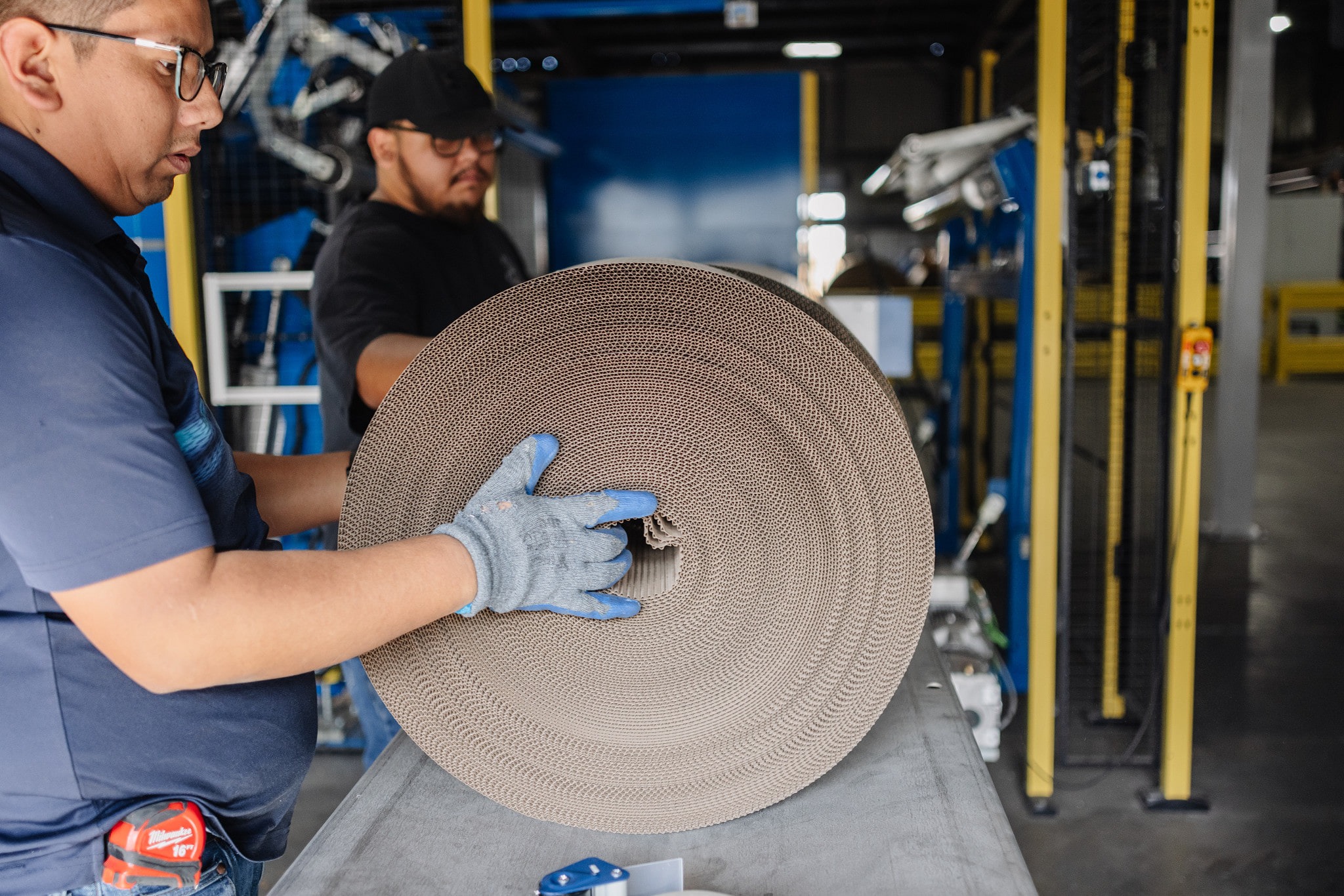 Two workers handle a large roll of material, preparing it for industrial use in a warehouse setting, emphasizing teamwork in a manufacturing process.